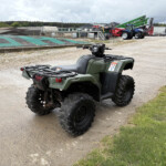 Green ATV in a gravel lot with heavy machinery and industrial buildings in the background.