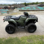 Green ATV parked on a gravel lot with industrial buildings and green fields in the background
