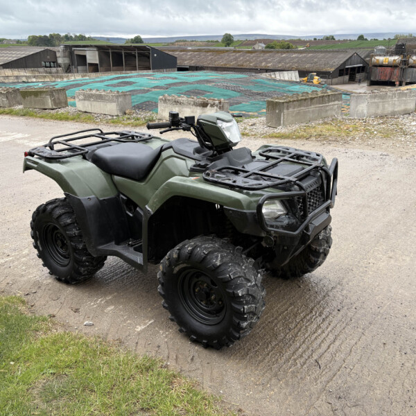 Green ATV parked on a gravel lot with farm buildings and fields in the distance under a cloudy sky