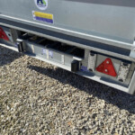 Rear view of a silver metal trailer on gravel, showing red taillight panels, bumper rails, and warning stickers on the back panel.