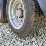 Close-up of a vehicle wheel with a steel rim, lug nuts, and a tire resting on gravel ground.