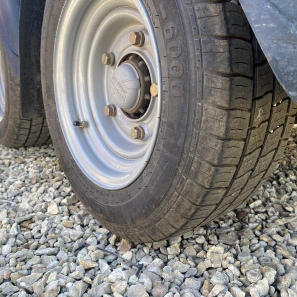 Close-up of a vehicle wheel with a steel rim, lug nuts, and a tire resting on gravel ground.