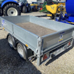 Open-top metal utility trailer on a gravel surface, with blue tractors and farming equipment in the background.