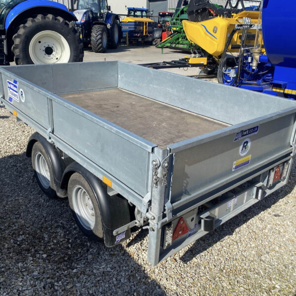 Open-top metal utility trailer on a gravel surface, with blue tractors and farming equipment in the background.