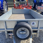 Metal utility trailer with a spare tire mounted on the side, parked among blue tractors and red farming equipment on a gravel lot.