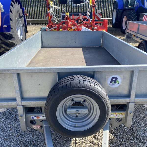 Metal utility trailer with a spare tire mounted on the side, parked among blue tractors and red farming equipment on a gravel lot.