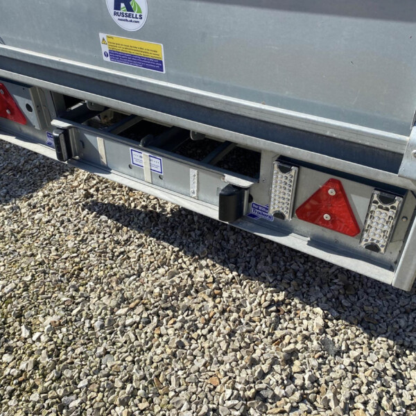 Rear view of a silver metal trailer on gravel, showing red taillight panels, bumper rails, and warning stickers on the back panel.
