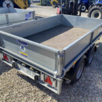 Metal utility trailer in a gravel lot, with stickers on the sides and other trailer equipment visible nearby; a blue tractor is in the background.