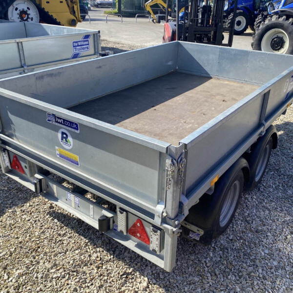Metal utility trailer in a gravel lot, with stickers on the sides and other trailer equipment visible nearby; a blue tractor is in the background.