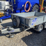 Blue industrial trailer with large wheel, hoses coiled on its side, and other yellow and red machinery in a yard; brand labels visible on the trailer.