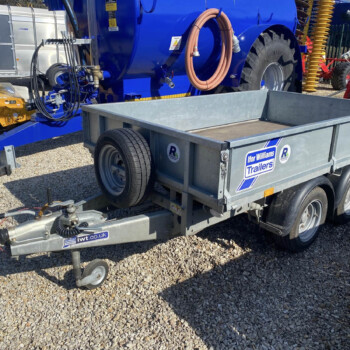 Blue industrial trailer with large wheel, hoses coiled on its side, and other yellow and red machinery in a yard; brand labels visible on the trailer.