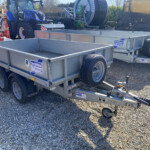 Silver metal trailer beds on a gravel lot, with a blue tractor and red machinery in the background; Trailers labeled 'Williams Trailers' are visible.