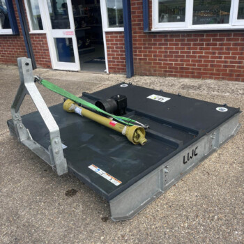 Outdoors, a flatbed lift platform with a green strap and yellow pole lies in front of a brick building with a glass door and windows, suggesting construction or maintenance equipment ready for use.