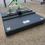 Industrial hydraulic lifting mechanism mounted on a dark gray metal platform, secured with bright green straps to a flat base on a paved surface