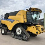 Yellow New Holland combine harvester with tracked wheels parked outdoors beside storage silos under a cloudy sky.