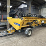 Yellow New Holland farming implement on a trailer inside a metal-walled workshop, with hoses and framework above it.