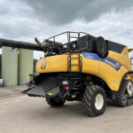 New Holland CR8.90 combine harvester with extended unloading auger in an industrial yard, front header visible and silos in background.