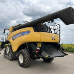 Yellow New Holland agricultural combine harvester with a long black unloading auger extended, parked on a paved area under a cloudy sky, showing its oversized tracked rear wheels and equipment.