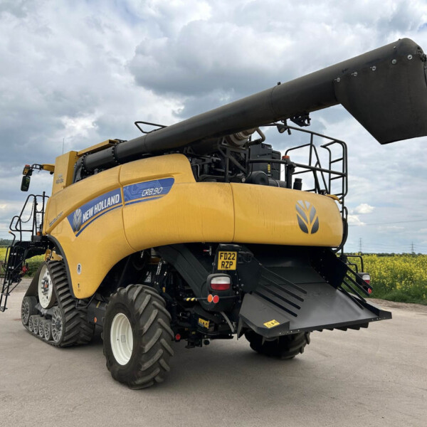 Yellow New Holland agricultural combine harvester with a long black unloading auger extended, parked on a paved area under a cloudy sky, showing its oversized tracked rear wheels and equipment.