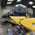 Yellow New Holland agricultural harvester with black mechanical arms inside a dusty shed, large machinery awaiting use in a barn-like workspace.