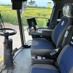 Interior of a tractor cab with a steering wheel, control panel and joystick, view of a yellow flower field outside through the windows