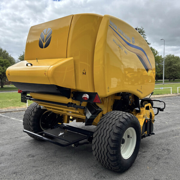 Bright yellow New Holland combine harvester parked on a paved lot, large rear wheels visible and a blue logo on the side, with trees and a cloudy sky in the background.