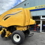 Yellow New Holland agricultural machine (Roll-Belt 150) parked outside a showroom with Russell’s sign in blue and yellow above the entrance, front-left view.