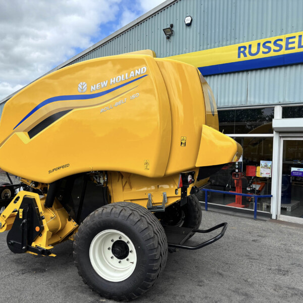 Yellow New Holland agricultural machine (Roll-Belt 150) parked outside a showroom with Russell’s sign in blue and yellow above the entrance, front-left view.