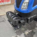 Close-up of a blue vehicle’s front end showing a heavy-duty hitch mechanism and coil springs on the ground near a brick wall sign.