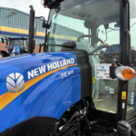 Close-up of a blue New Holland tractor cabin at a dealership, with branding on the side and signs in the background reading Holland and Russells.