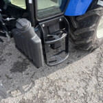 Close-up of a heavy-duty truck's front area showing the diesel fuel cap, hoses, and a large tire on a gravel surface