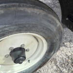 Close-up of a weathered tire mounted on a white steel wheel with five lug bolts and a black center cap, on a gravel surface.