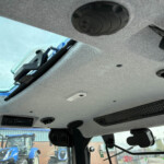 Interior view of a vehicle cab ceiling with vents and overhead hardware; blue tractors sit outside through the windshield.