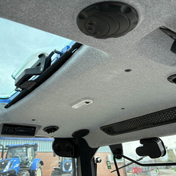 Interior view of a vehicle cab ceiling with vents and overhead hardware; blue tractors sit outside through the windshield.