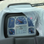 Close-up of a vehicle instrument cluster with RPM gauge, digital readouts, and warning icons under a transparent plastic cover