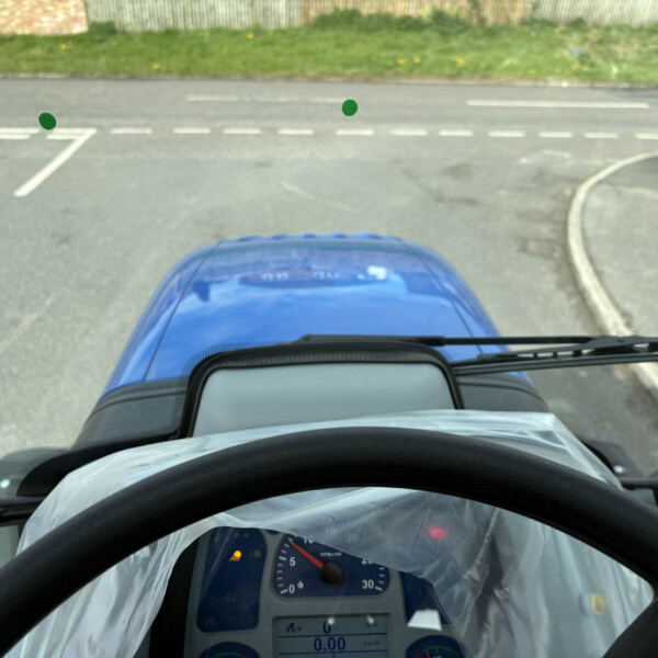 Driver's viewpoint from inside a blue car, looking at a road with dashed markings and a grassy verge ahead; the dashboard is in the foreground.