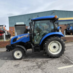 Blue New Holland tractors parked outside a workshop, with a red Manitou forklift visible in the background and a brick building.