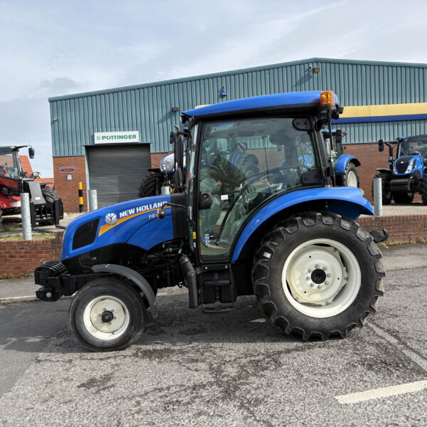 Blue New Holland tractors parked outside a workshop, with a red Manitou forklift visible in the background and a brick building.