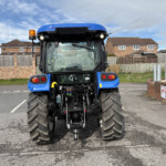 Rear view of a blue Manitou tractor/telehandler with large, rugged rear tires in a parking lot, houses and a wooden fence behind it.