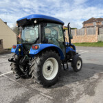 Blue tractor parked in a paved lot with brick walls and houses in the background.