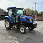 Blue New Holland tractor parked in a paved lot with houses and a brick wall in the background, front-right view.