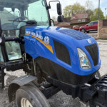Blue New Holland tractor front end close-up, showing the cab, headlights, and bold logo on a bright blue body.