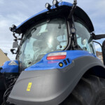 Close-up of a blue tractor cab with headlights and windshield, showing front fender and large tire visible nearby in a sunny outdoor setting.