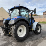 Blue agricultural tractor parked on a suburban street with brick walls and houses in the background under a cloudy sky in daylight.