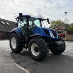 Blue New Holland tractor parked in a paved lot with brick houses and a cloudy sky behind it.