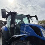 Close-up of a blue New Holland tractor cab with large side mirrors and dual roof lights, front grille visible in a parking lot.
