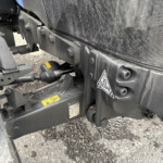 Close-up of a heavy-duty trailer chassis with bolts, brackets, and hoses amid a mesh panel and dark metal frames.