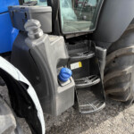 Close-up of a blue construction vehicle front, showing the cab steps, hydraulic connection, and a large tire nearby with dirt and grime on the body.