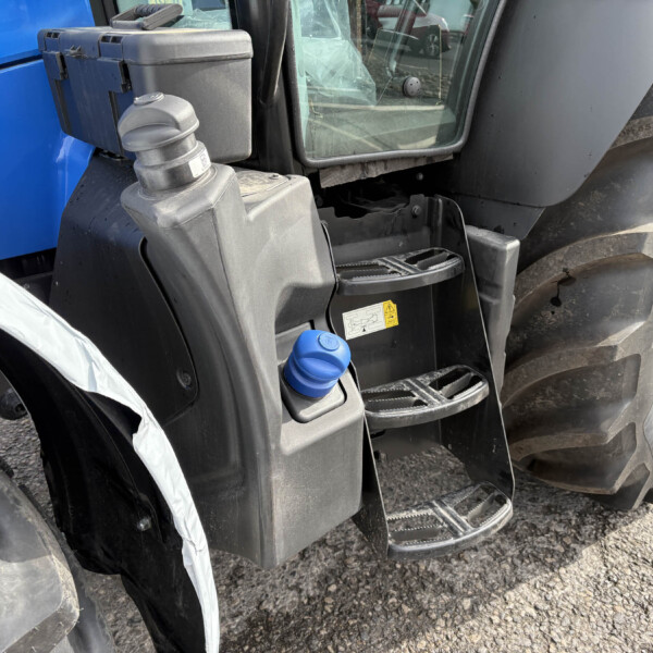 Close-up of a blue construction vehicle front, showing the cab steps, hydraulic connection, and a large tire nearby with dirt and grime on the body.