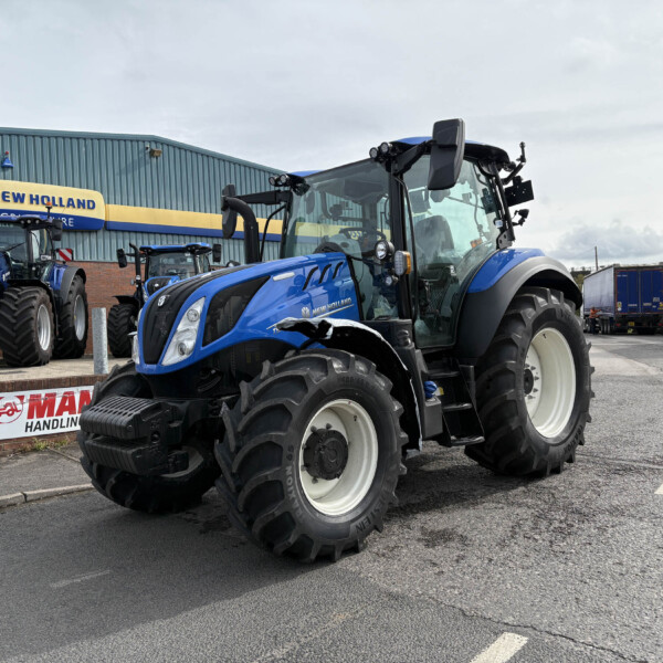 Blue New Holland tractors parked outside a dealership, with a large foreground tractor and a sign reading 'New Holland' on the building behind.